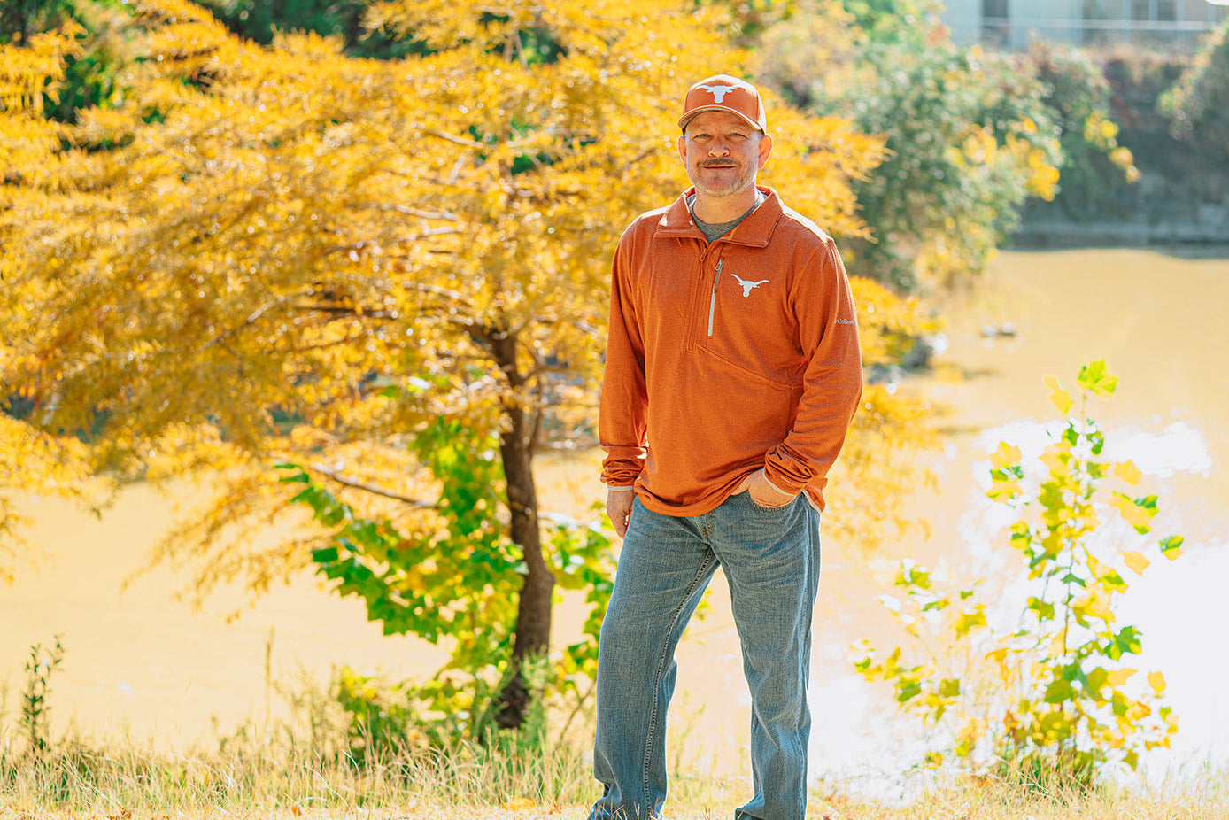 Man in an orange sweatshirt and cap standing in a park with trees and water in the background