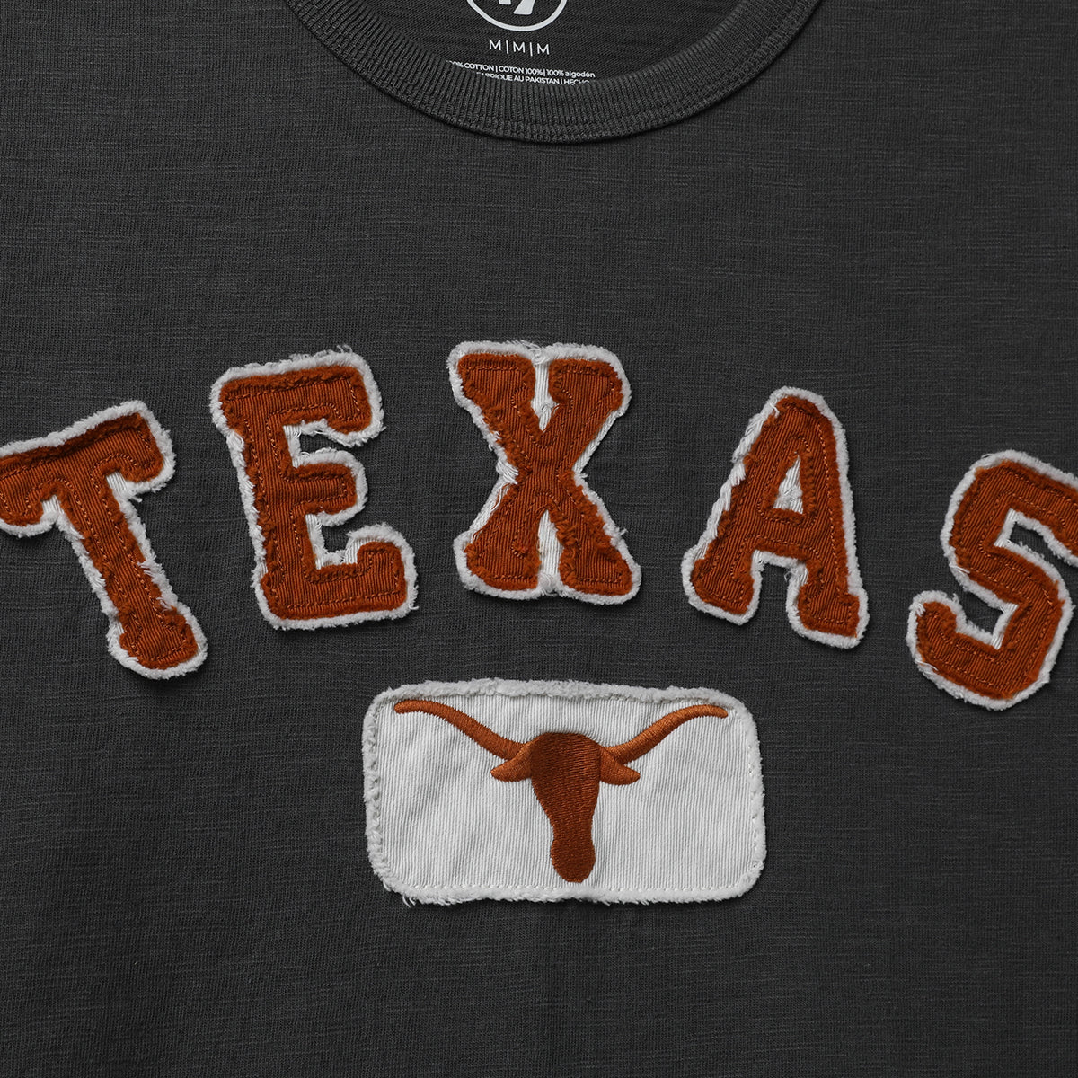 Close-up of a dark charcoal gray t-shirt featuring a textured brown and white embroidered 'TEXAS' graphic and a matching embroidered Longhorn patch below.