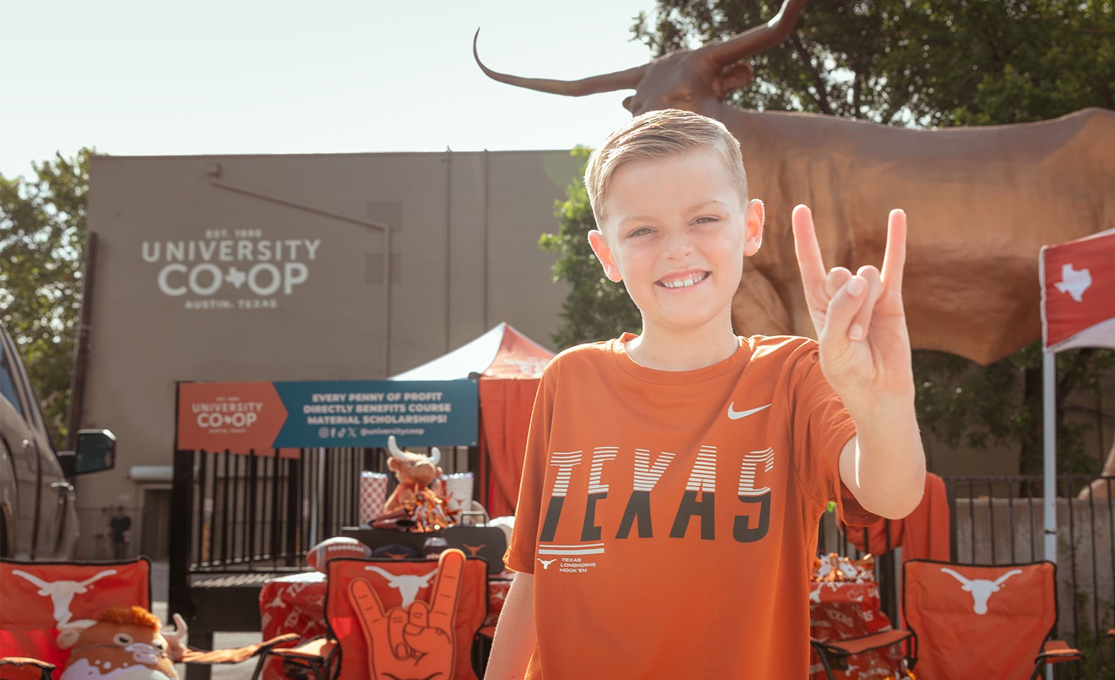A young boy smiling and doing a hook em sign, wearing an orange Nike Texas athletic t-shirt in an outdoor event setting with University Coop branding and a longhorn statue in the background.