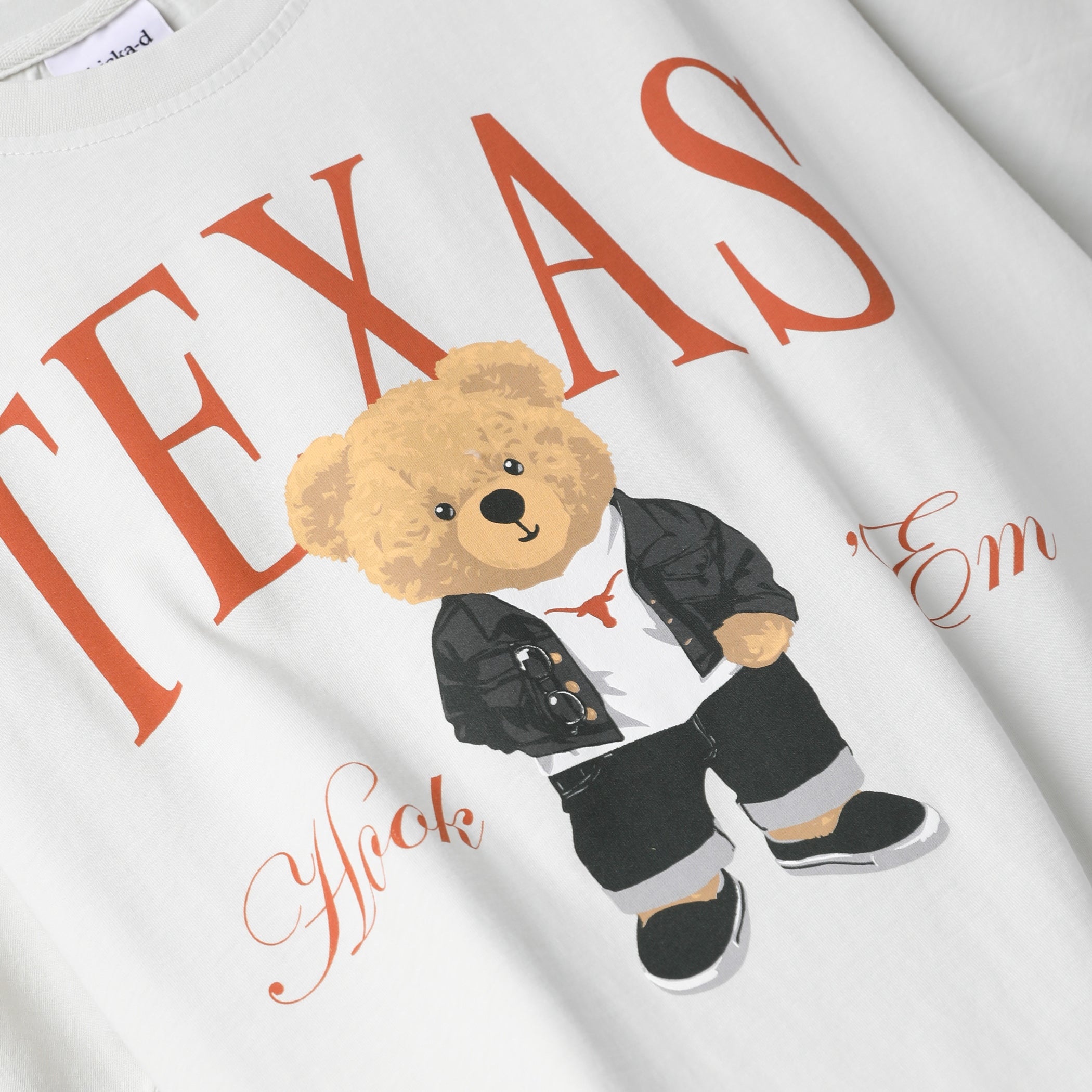 White graphic t-shirt featuring a cartoon teddy bear dressed in a black jacket and pants with a small Texas longhorn logo on its shirt. Large orange text reads 'TEXAS' across the chest, with additional orange script text below.