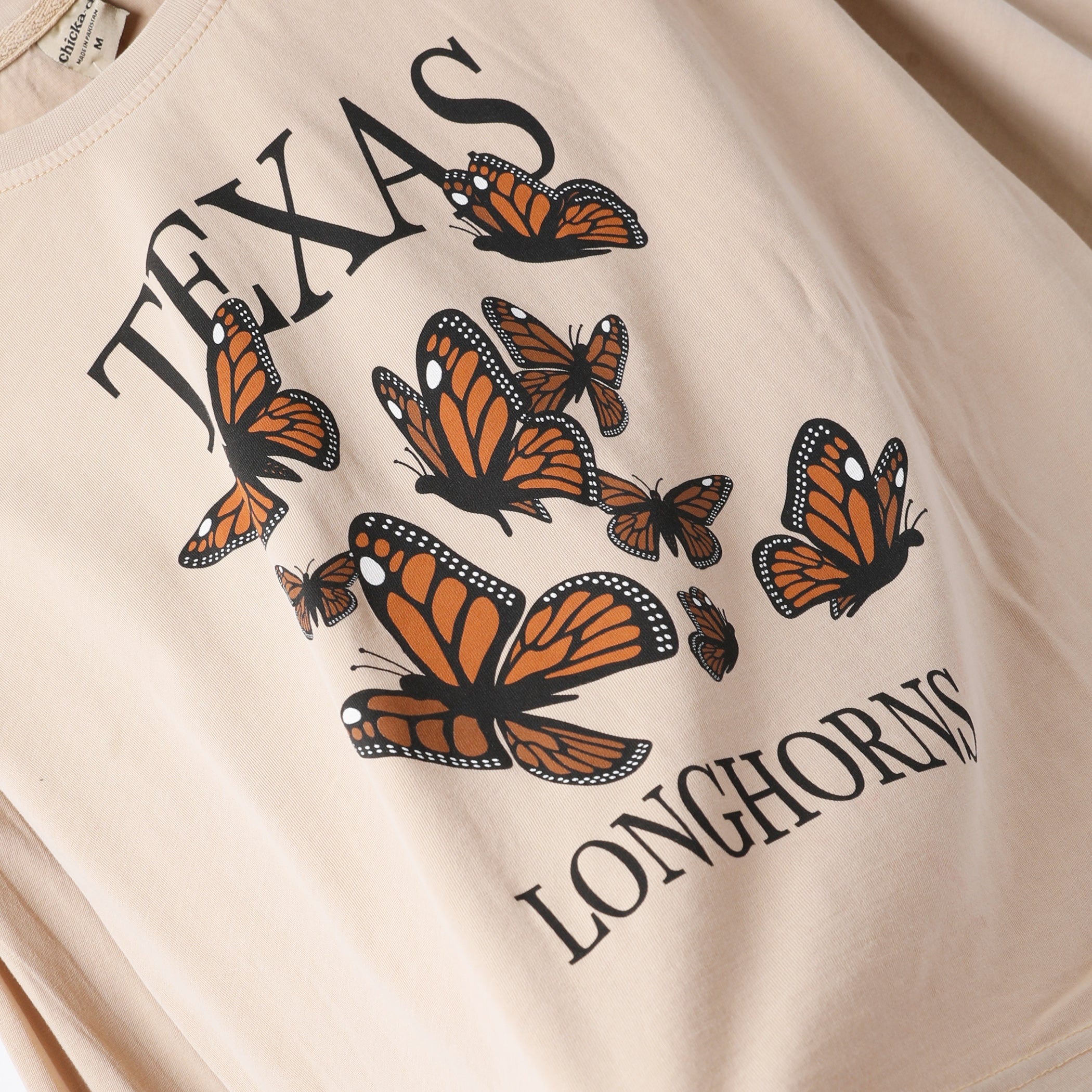 A close-up of a cream-colored t-shirt featuring a graphic print of orange and black monarch butterflies with the text 'TEXAS LONGHORNS' in black.