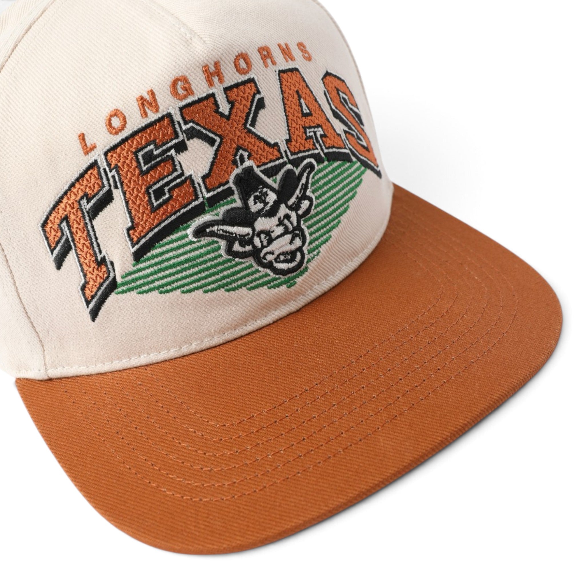A close-up of a Texas Longhorns baseball cap featuring a cream crown with orange and green embroidered lettering and a horned mascot, paired with a rust-orange flat brim.