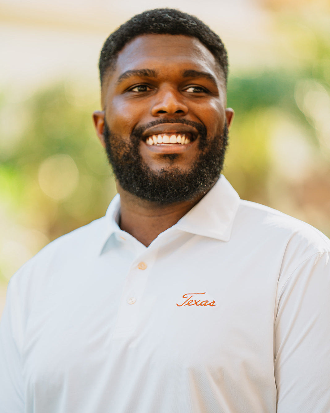 Man wearing a white Peter Millar polo shirt with 'Texas' embroidery against a blurred natural background