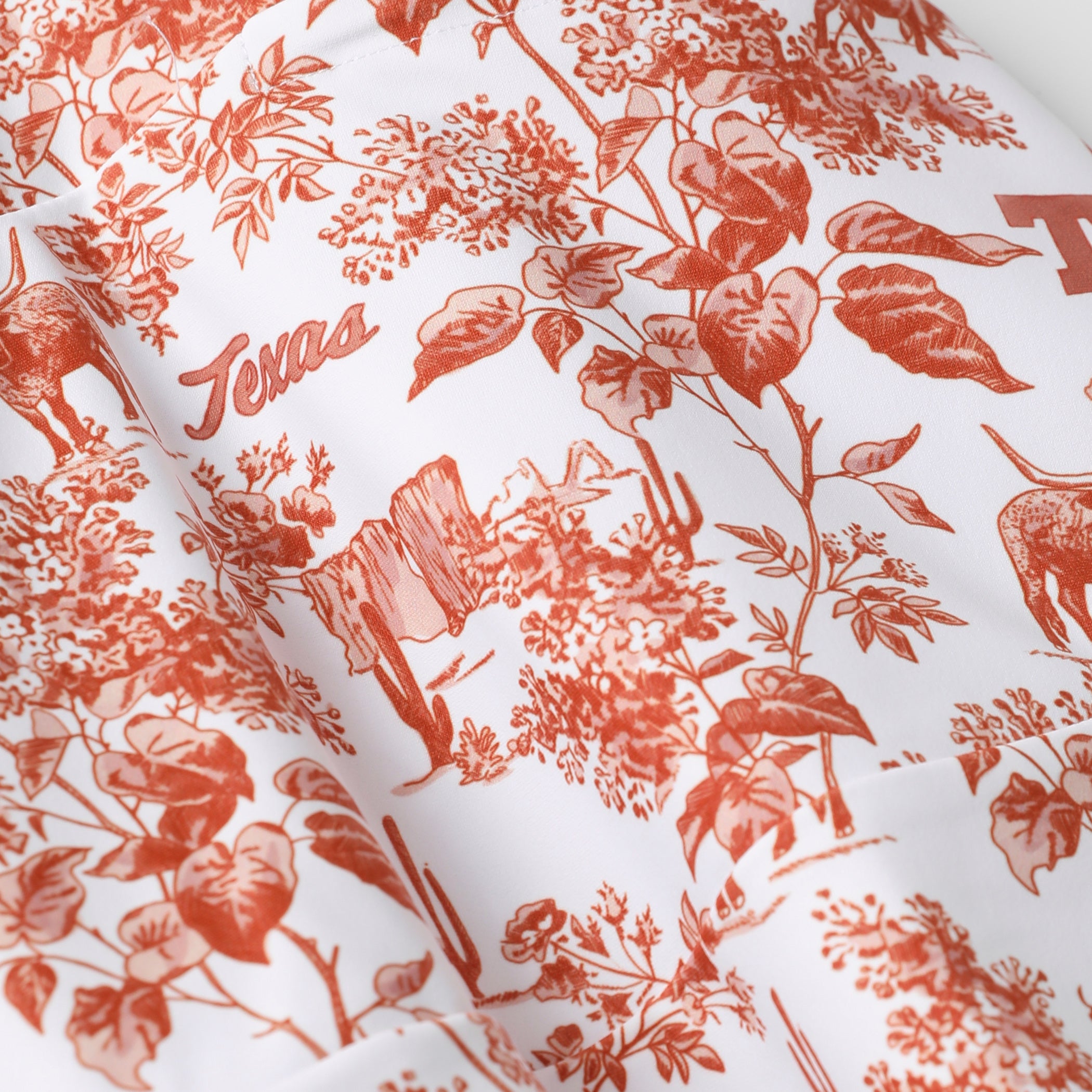 White fabric with a terracotta red Texas-themed toile pattern featuring cacti, flowers, and the word 'Texas'.