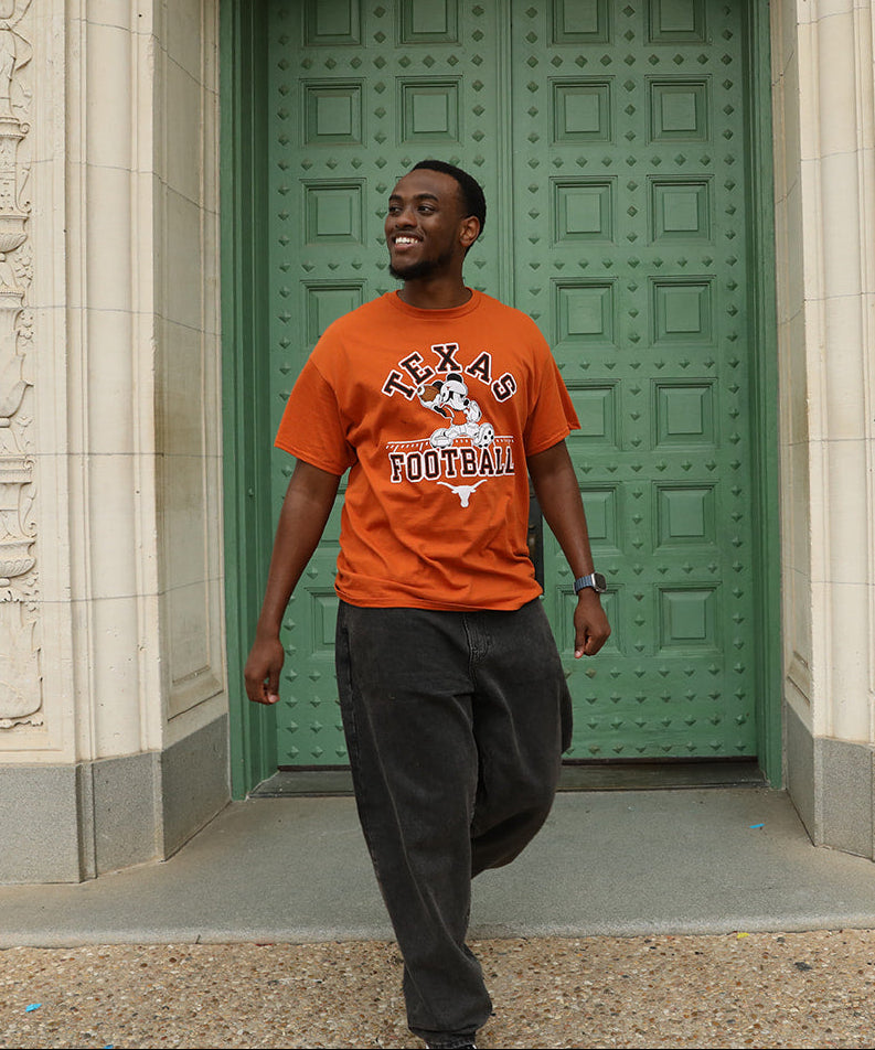 A man wearing an orange crewneck t-shirt with a 'Texas Football' graphic and dark gray sweatpants walking in front of a large green door.