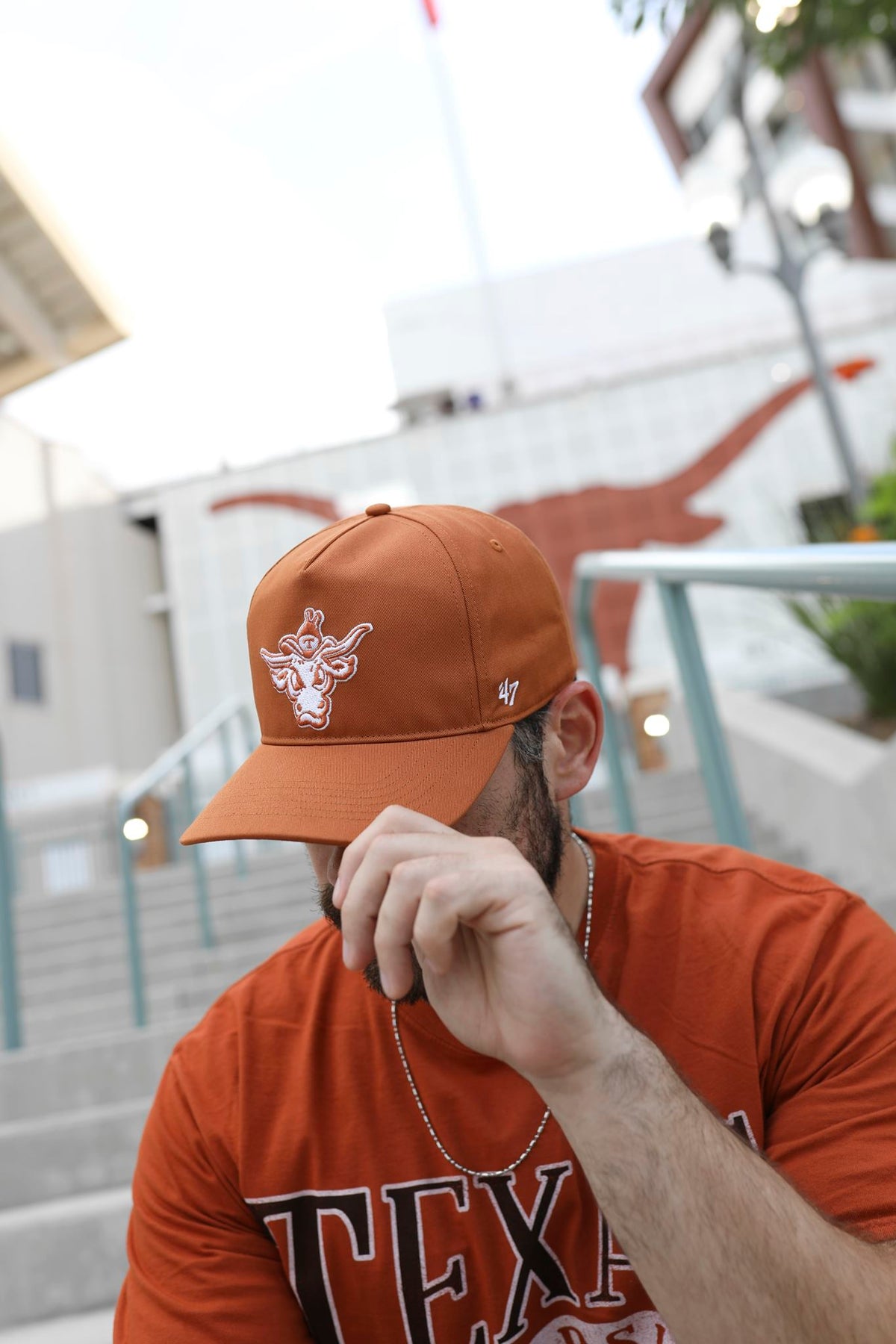 A man wearing a burnt orange Texas Longhorns baseball cap and matching burnt orange t-shirt, sitting outdoors in front of the Texas Longhorns stadium with the team's longhorn logo visible in the background.