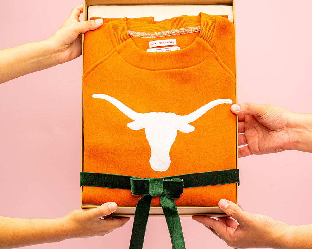 Orange shirt with a longhorn logo held by two hands against a pink background