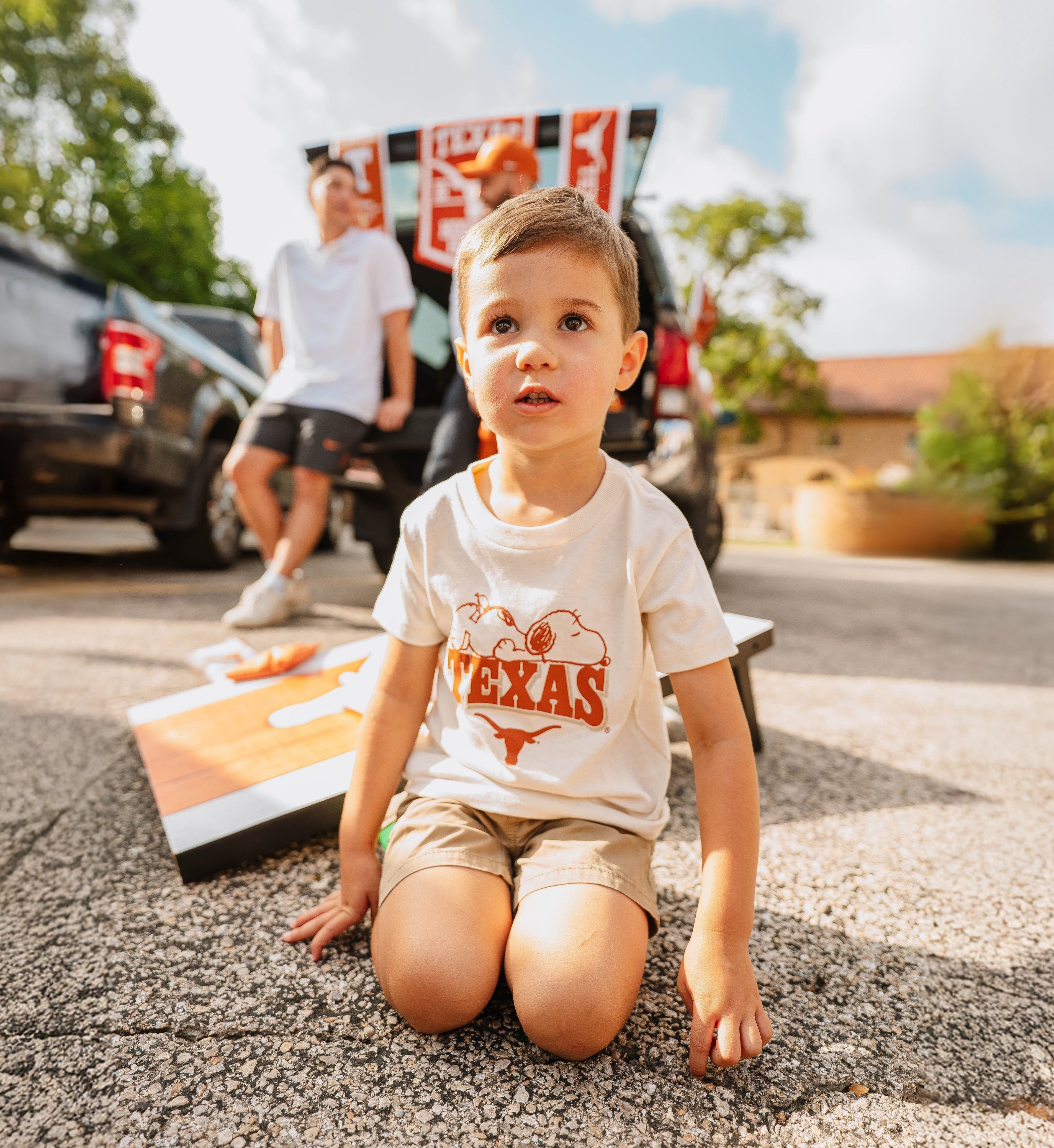 Child wearing a 'Texas' shirt sitting on the ground with a car and person in the background. Texas longhorn merch