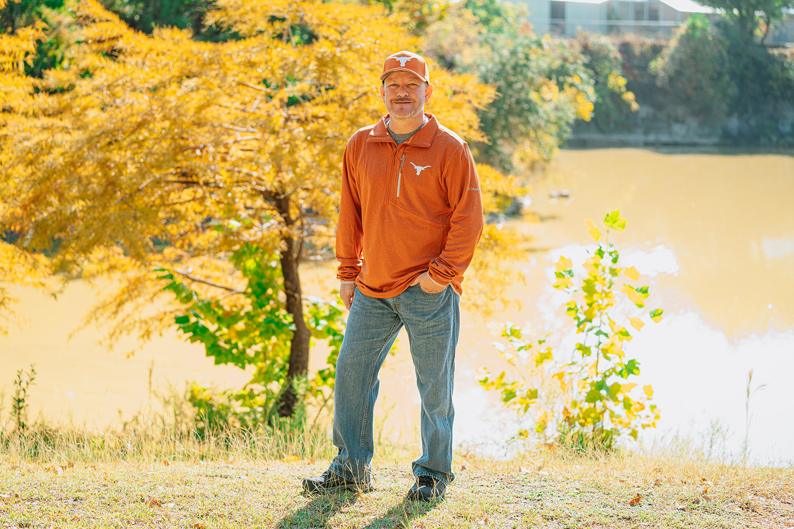 Man in an orange sweatshirt and cap standing in a park with trees and water in the background