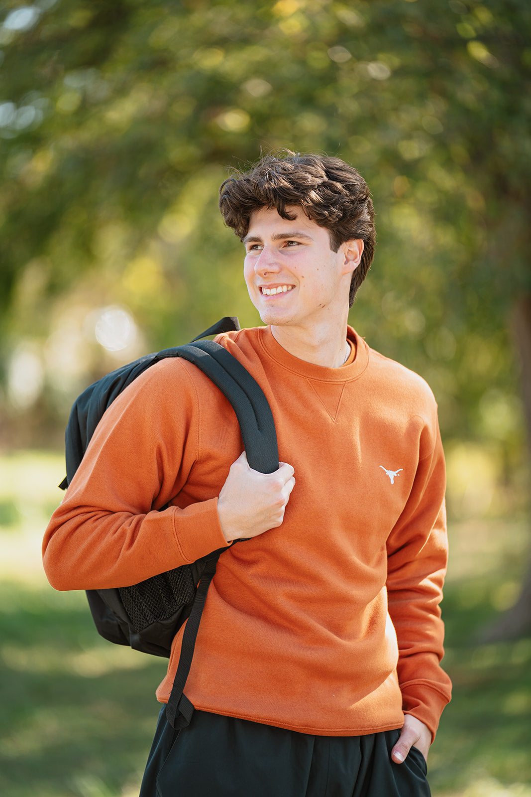 Man wearing an orange sweatshirt with a logo, holding a black backpack outdoors.