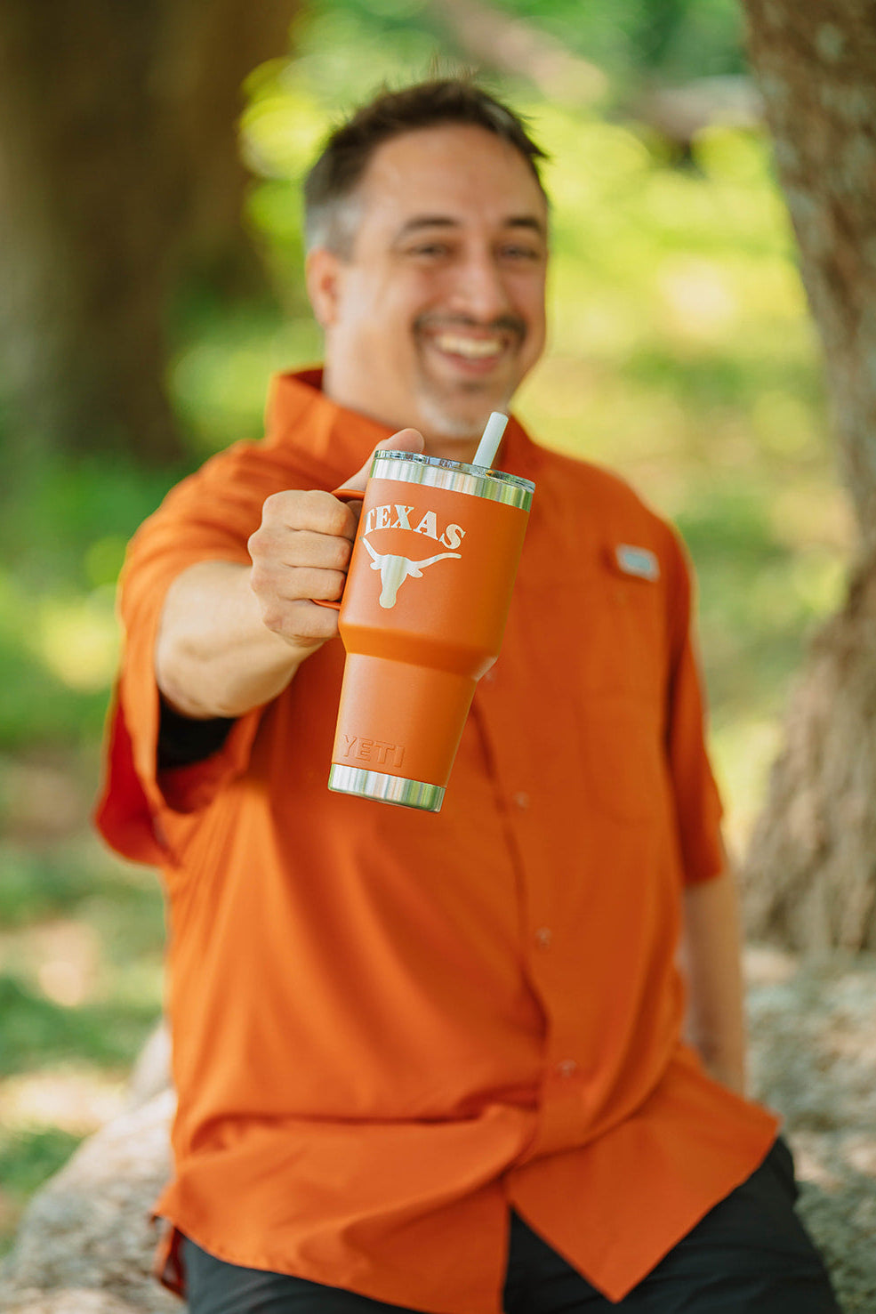 A man in an orange button-down shirt holding up a stainless steel tumbler with the Texas Longhorns logo and 'TEXAS' text, set against a blurred outdoor background of trees and foliage.