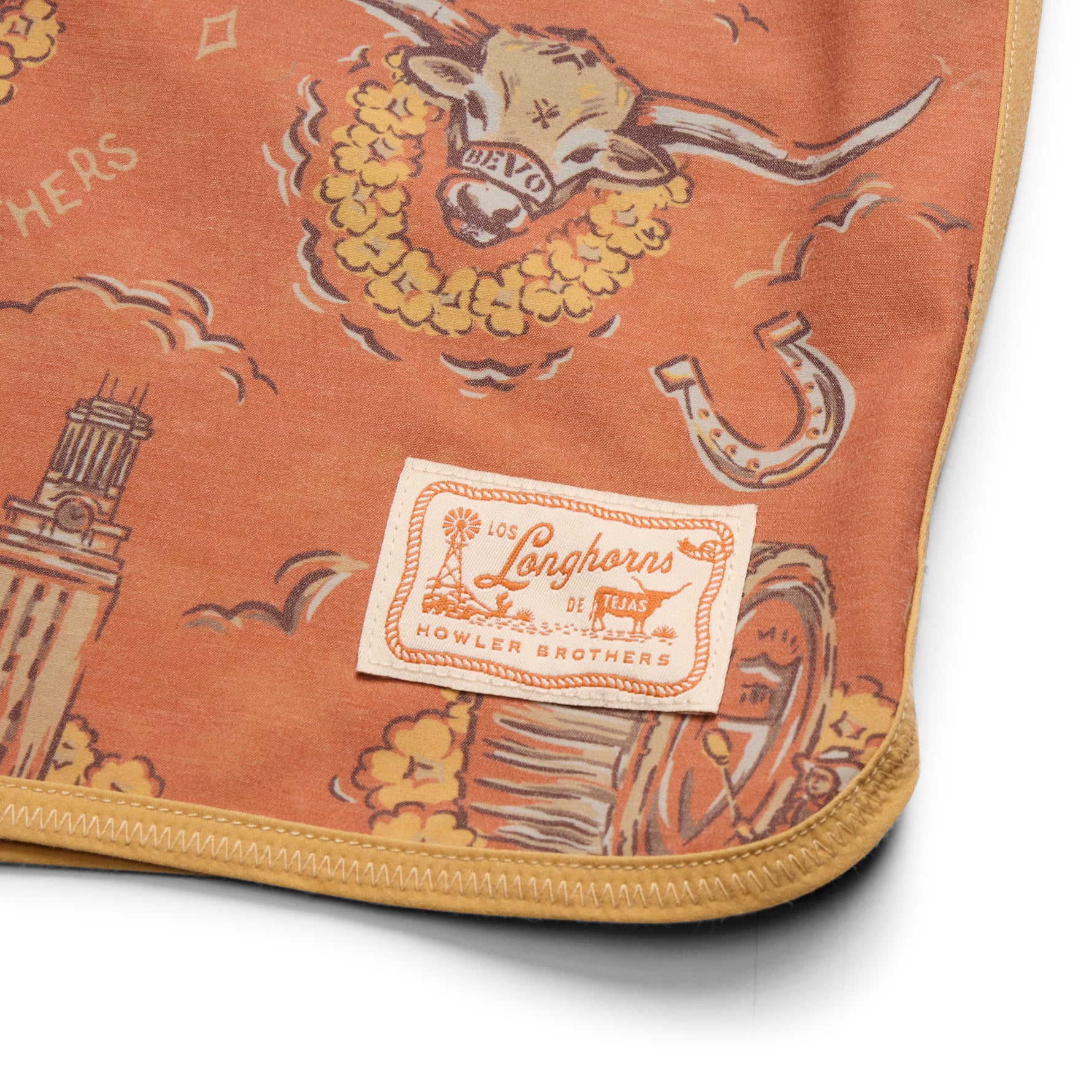 A close-up of a folded terracotta orange blanket featuring a western-themed print with a steer, horseshoe, and a sewn-on label that reads 'Los Longhorns de Texas' and 'Howler Brothers'.