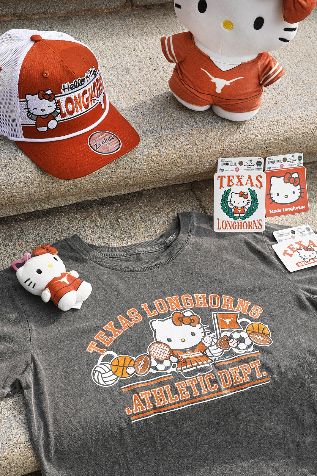 A Texas Longhorns Hello Kitty themed apparel flat-lay featuring a dark gray graphic t-shirt, an orange and white trucker hat, a Hello Kitty plush doll, and two packaged stickers, all arranged on a stone surface.