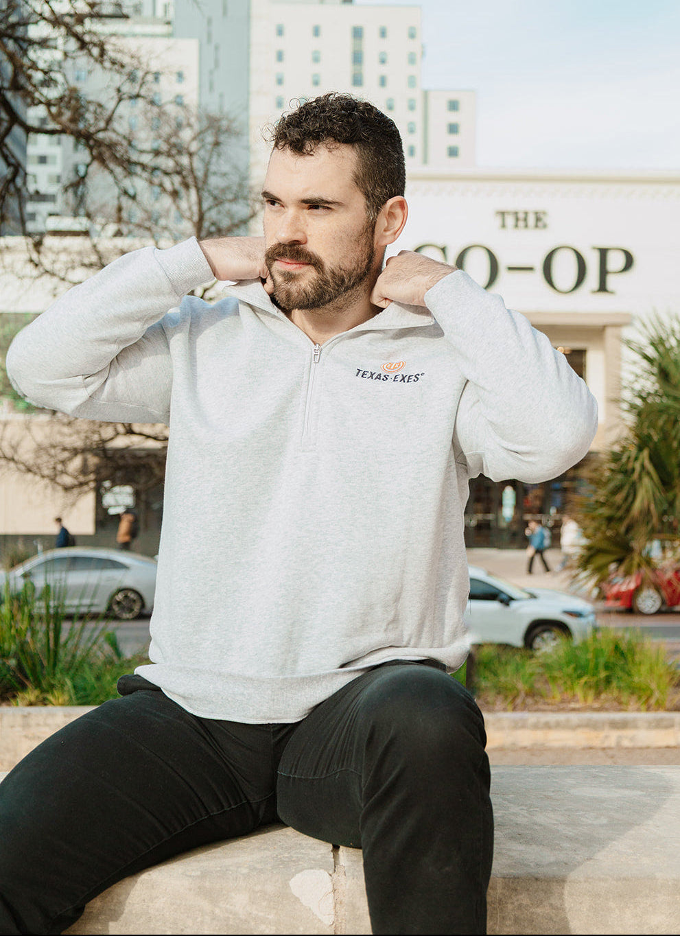 Man sitting outdoors wearing a white hoodie with a logo, in front of a building with 'The Co-op' sign.