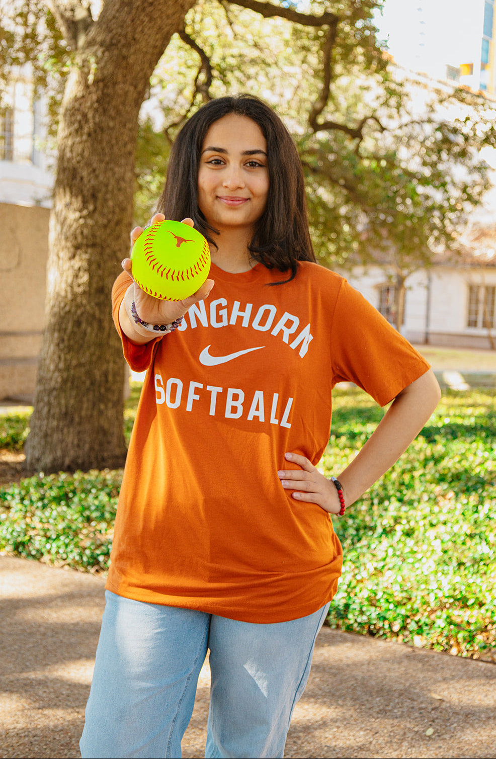 Person wearing an orange Longhorn Softball shirt holding a yellow softball outdoors.