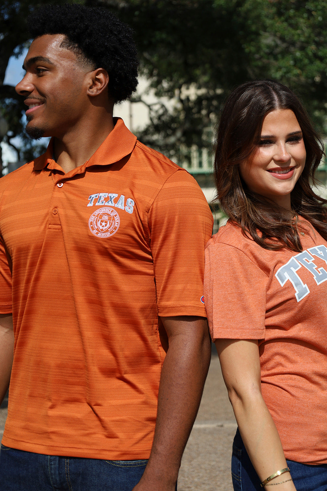 Two students wearing orange Texas athletic shirts walking outdoors. The male model wears a long-sleeve polo shirt with a circular Texas logo on the left chest, and the female model wears a short-sleeve crewneck shirt with 'TEXAS' lettering on the front.