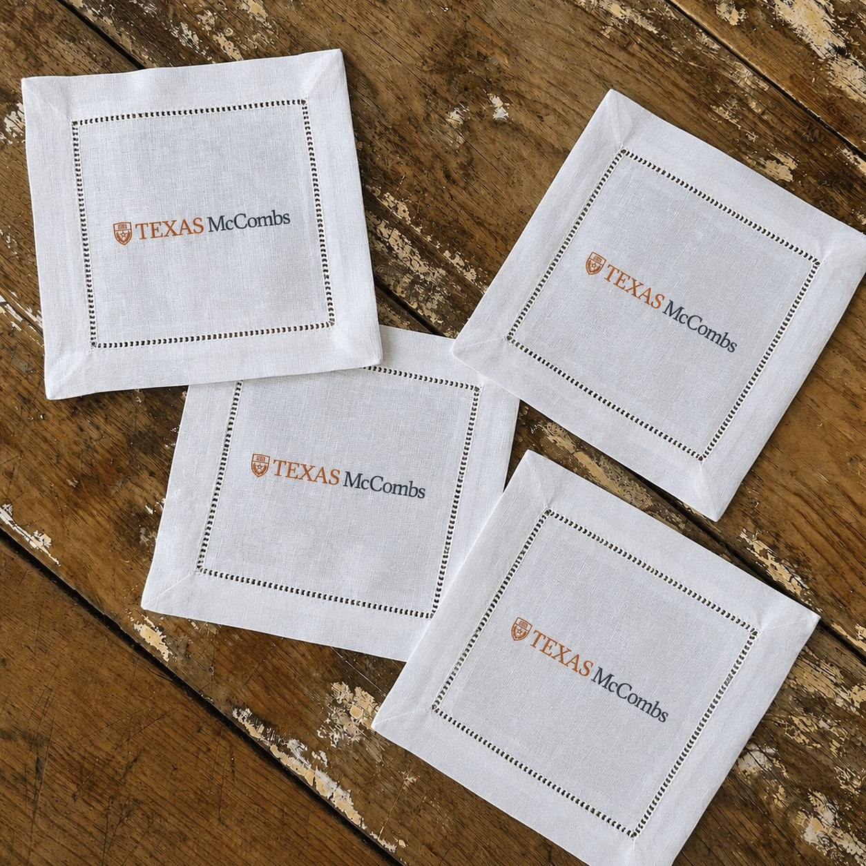 Four white square paper napkins with stitched borders and the printed logo 'TEXAS McCombs' arranged on a rustic wooden tabletop.
