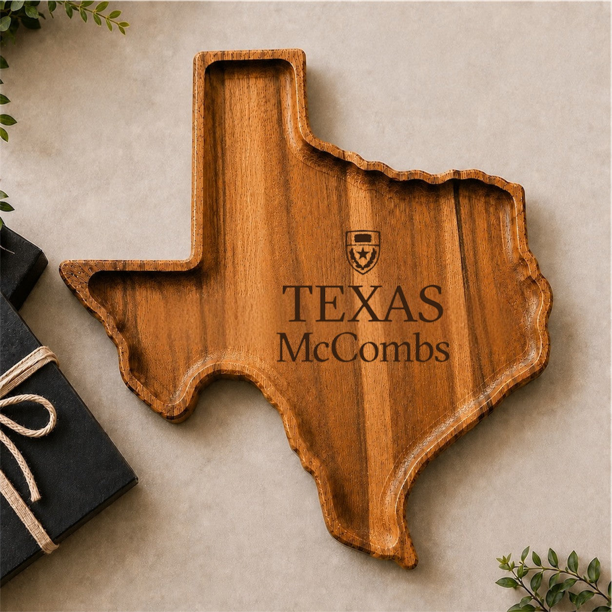 A wooden serving tray shaped like the state of Texas, engraved with a shield logo and the text 'TEXAS McCombs', displayed on a neutral beige surface with decorative greenery and a gift box at the side.