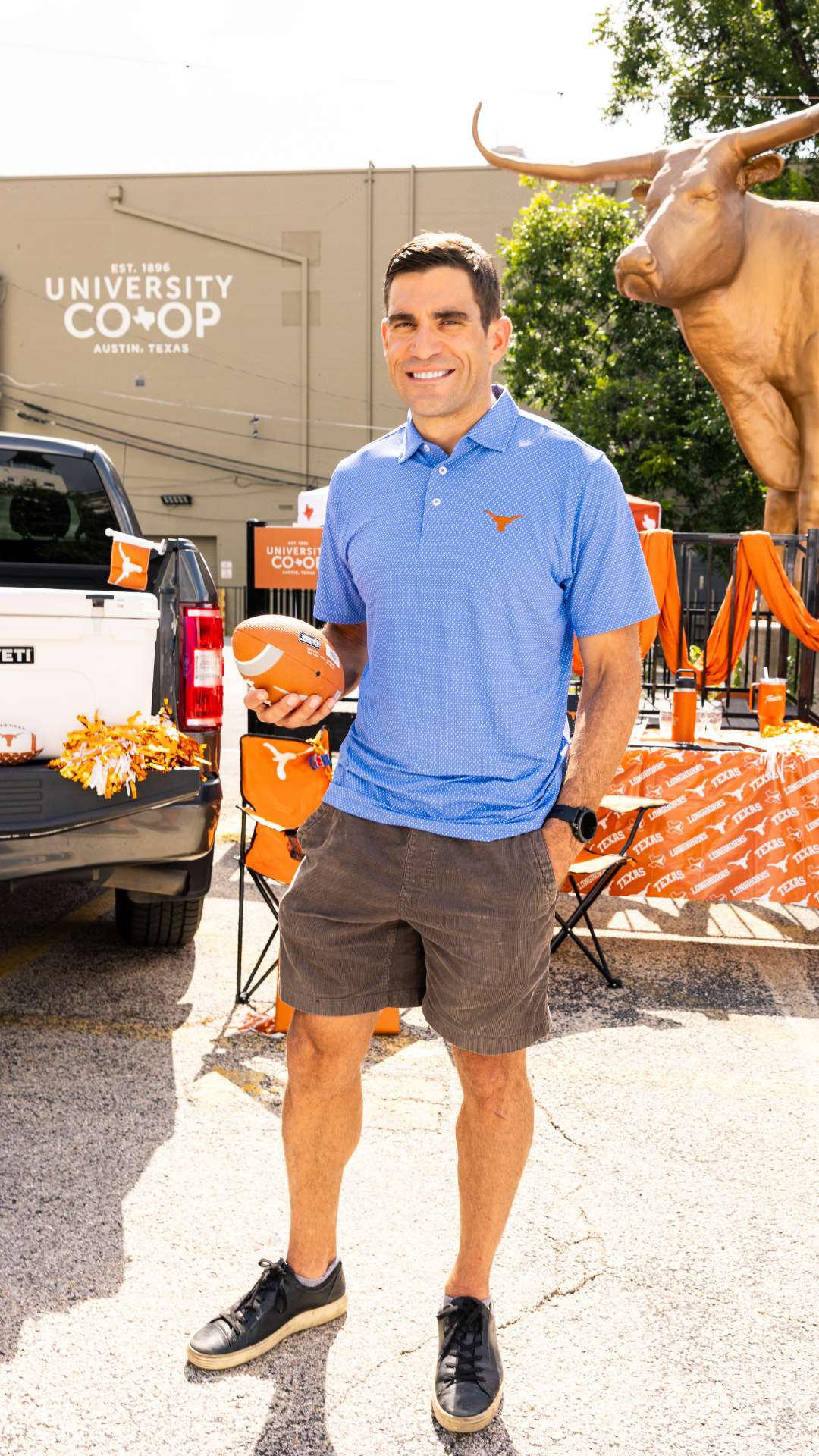 A man wearing a blue polo shirt, brown shorts, and black sneakers stands outdoors holding a football. He is in front of a building with 'University Coop' signage, a large longhorn statue, and orange University-branded event decorations.