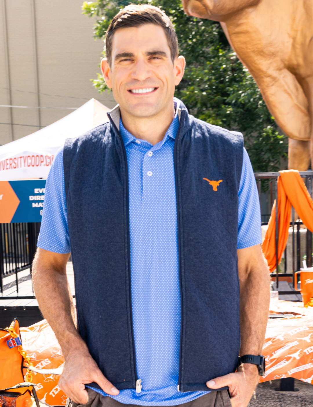 A man wearing a navy blue zip-up vest with a small orange Texas Longhorns logo over a light blue polo shirt, standing outdoors in front of a large bronze bull statue and orange event chairs.