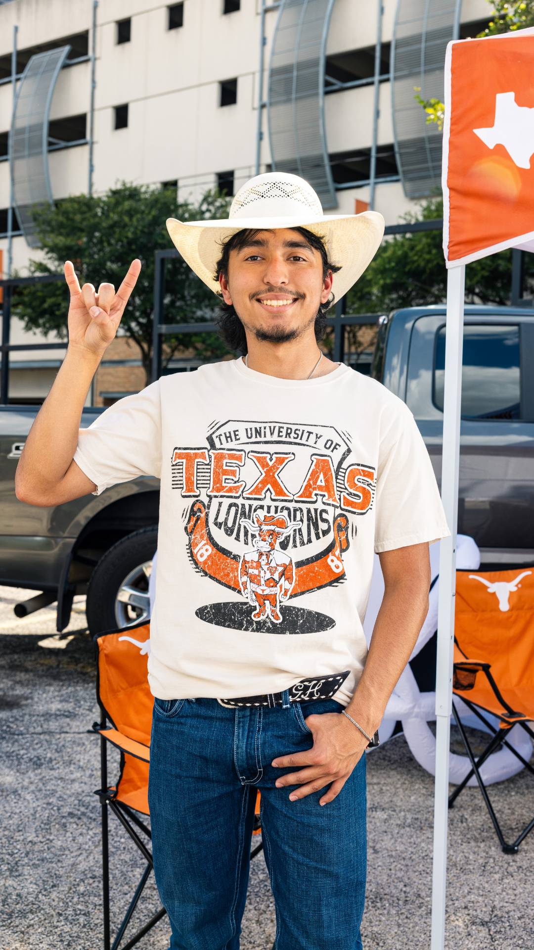 A man wearing a white cowboy hat and a white graphic t-shirt that reads 'The University of Texas Longhorns' stands outdoors in front of a building and truck, making a hand gesture. An orange Texas flag and folding chairs are visible in the background.