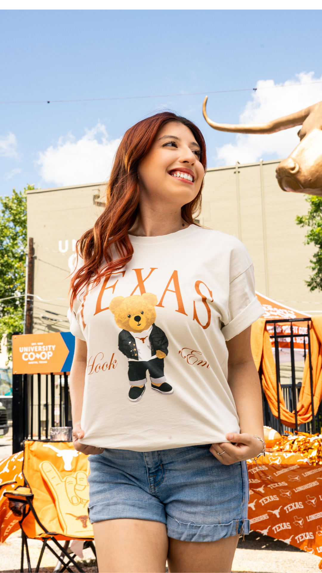 A woman wearing a white Texas-themed graphic t-shirt with a teddy bear and 'Texas Longhorns' design, paired with denim shorts, standing outdoors in front of a building with a longhorn statue.
