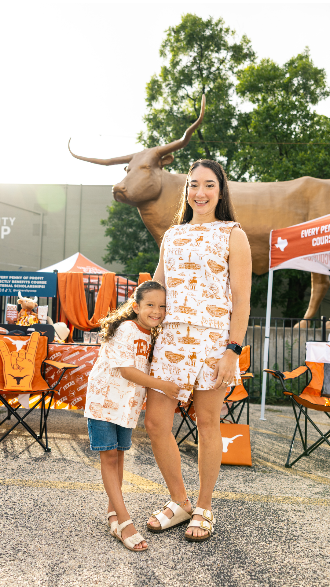 A woman and a young girl wearing matching Texas-themed outfits with an orange and white hot air balloon print, standing in front of a large Texas longhorn statue at an outdoor event.