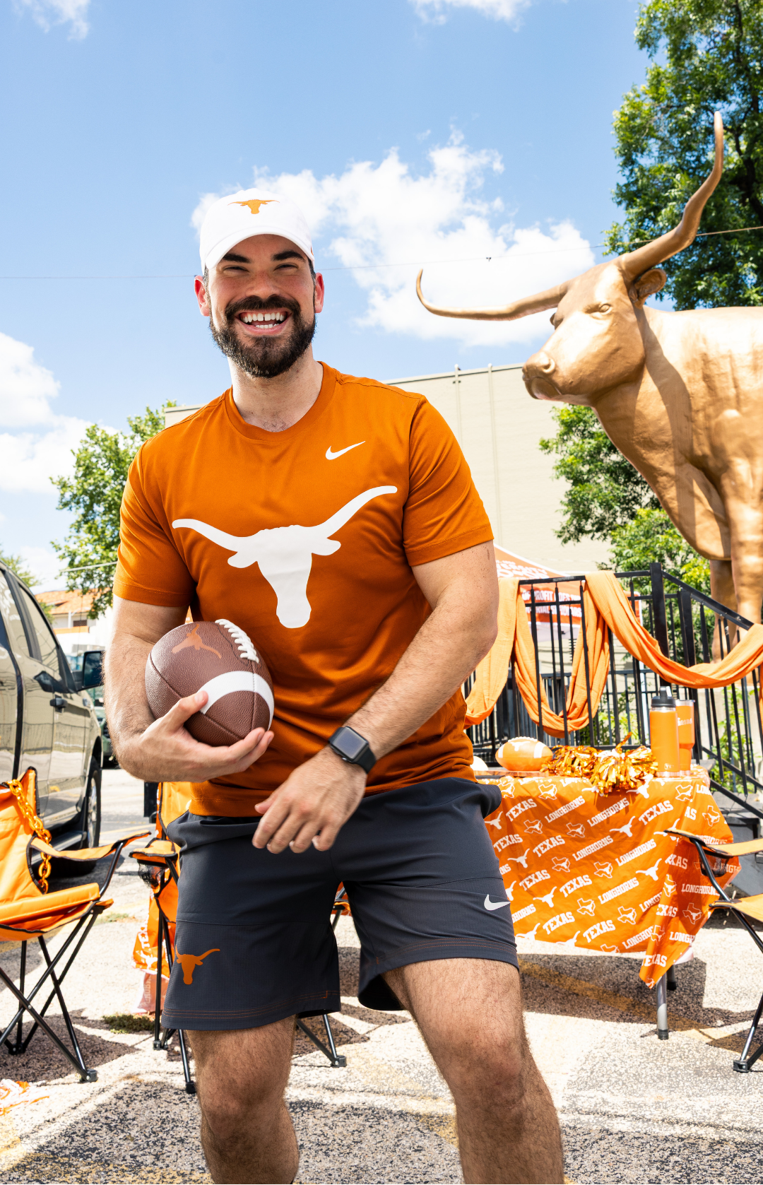 A smiling man wearing a burnt orange Texas Longhorns athletic t-shirt and matching shorts stands outdoors holding a football. The shirt features a white Longhorns logo and a Nike swoosh, with the Texas Longhorns brand also visible on the shorts.