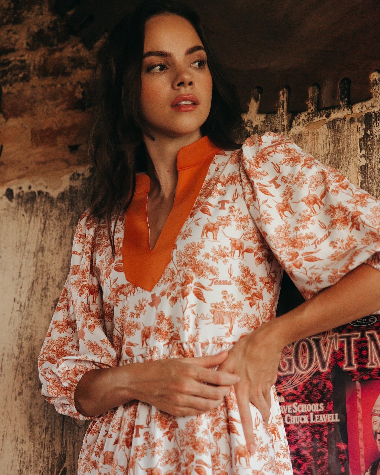 Woman wearing a white and burnt orange floral dress with an orange top against a textured wall.