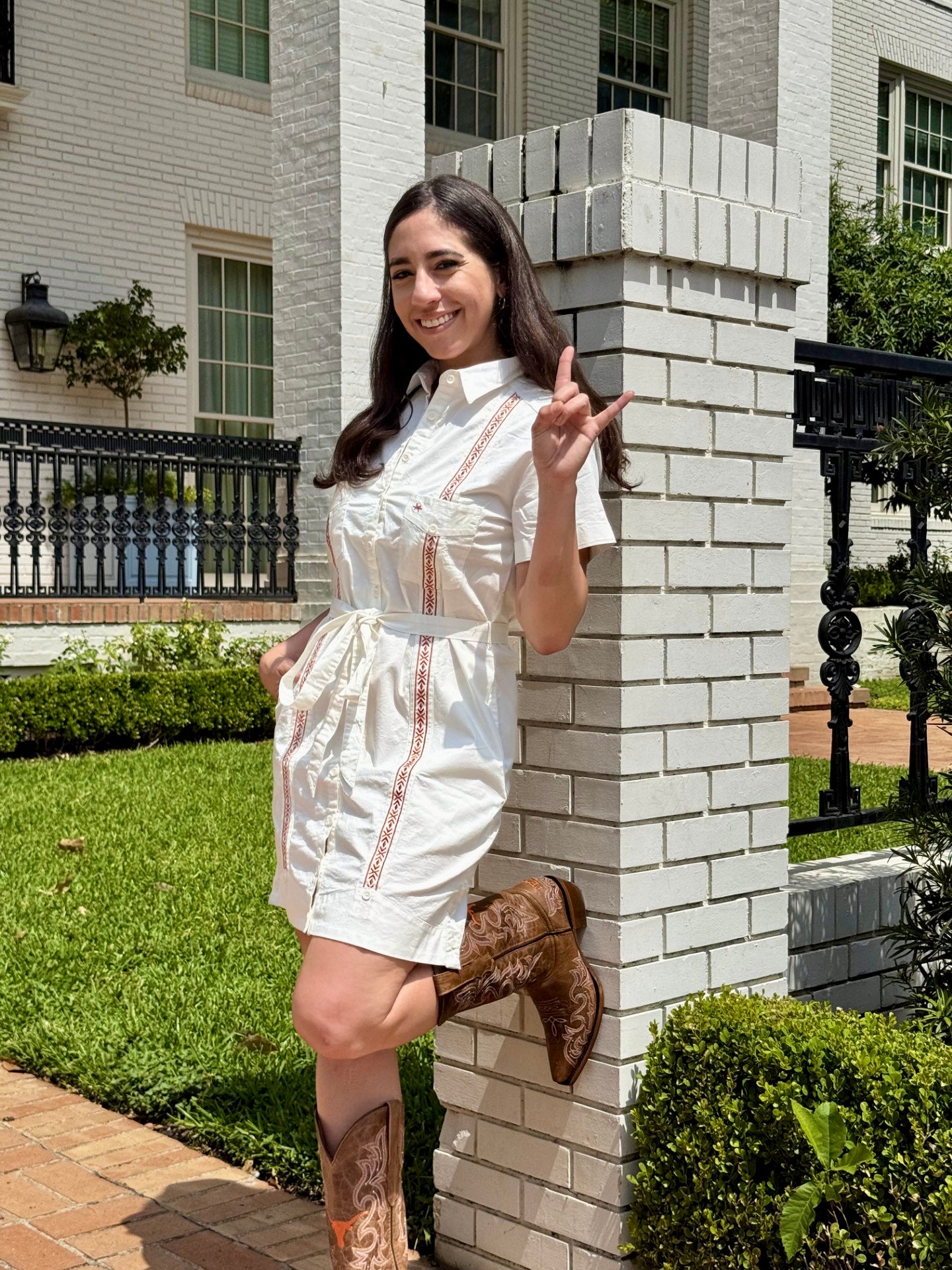 woman wearing white collared dress with tie front, burnt orange stitching detail and cowboy boots